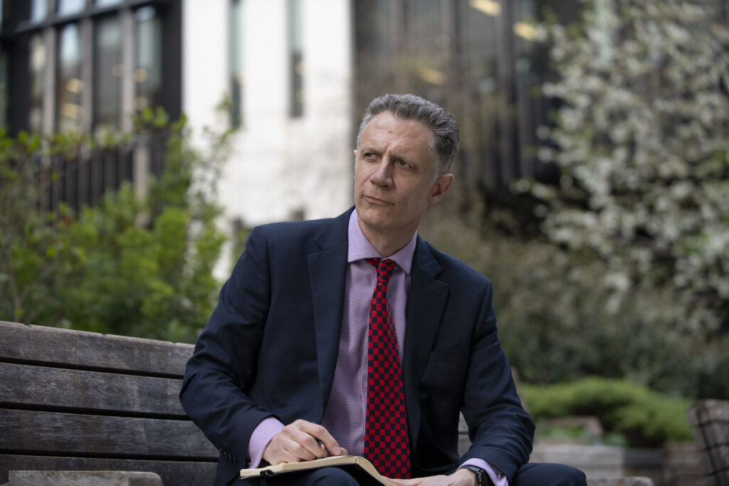 Photo of a mentor in a business suit, surounded by tall buildings, sitting on a park bench, thinking and making notes, about Mentoring Discovery Calls.